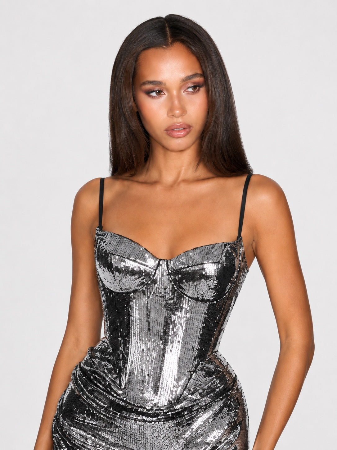 A woman modeling a silver sequined corset-style mini dress with thin black spaghetti straps against a light gray studio background.