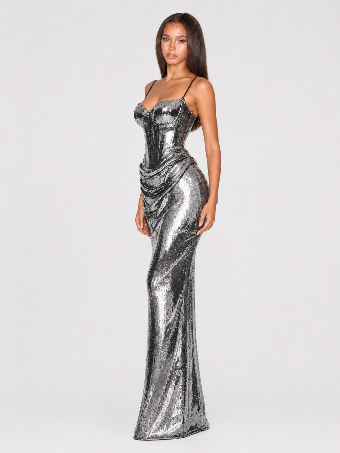 A woman modeling a floor-length metallic silver sequin gown with thin spaghetti straps, a sweetheart neckline, and a ruched corset-style bodice against a light gray studio background.