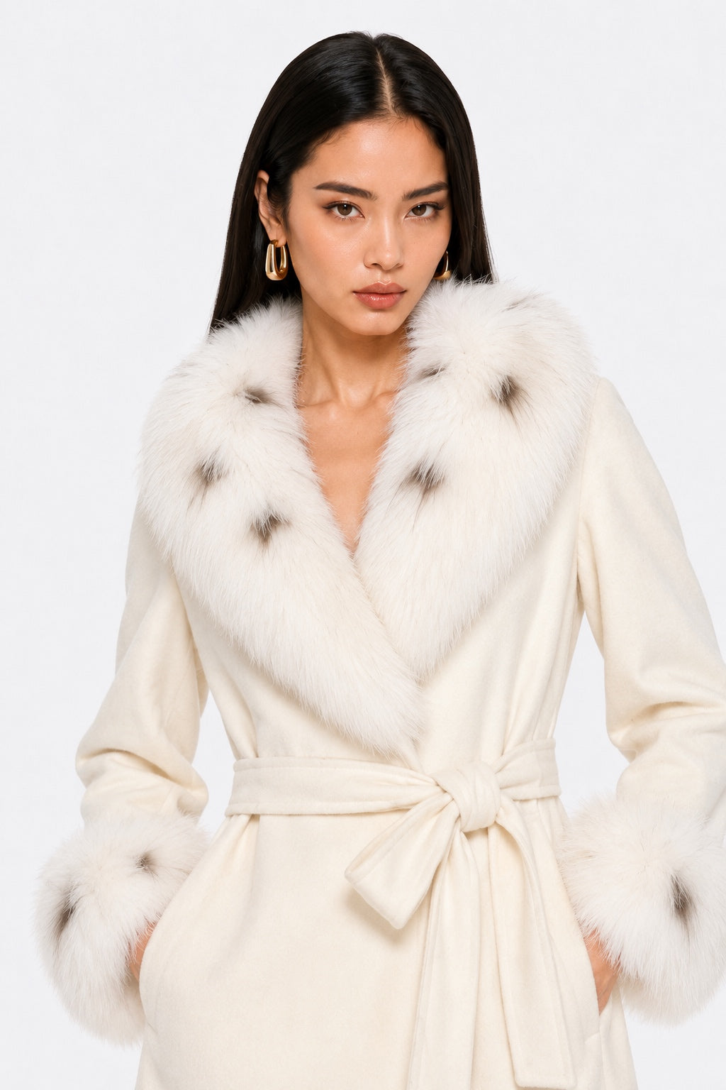 A woman wearing a cream-colored wrap coat with a large white fur collar featuring dark brown tips, a matching waist tie belt, and fluffy cuffs, set against a plain white studio background.