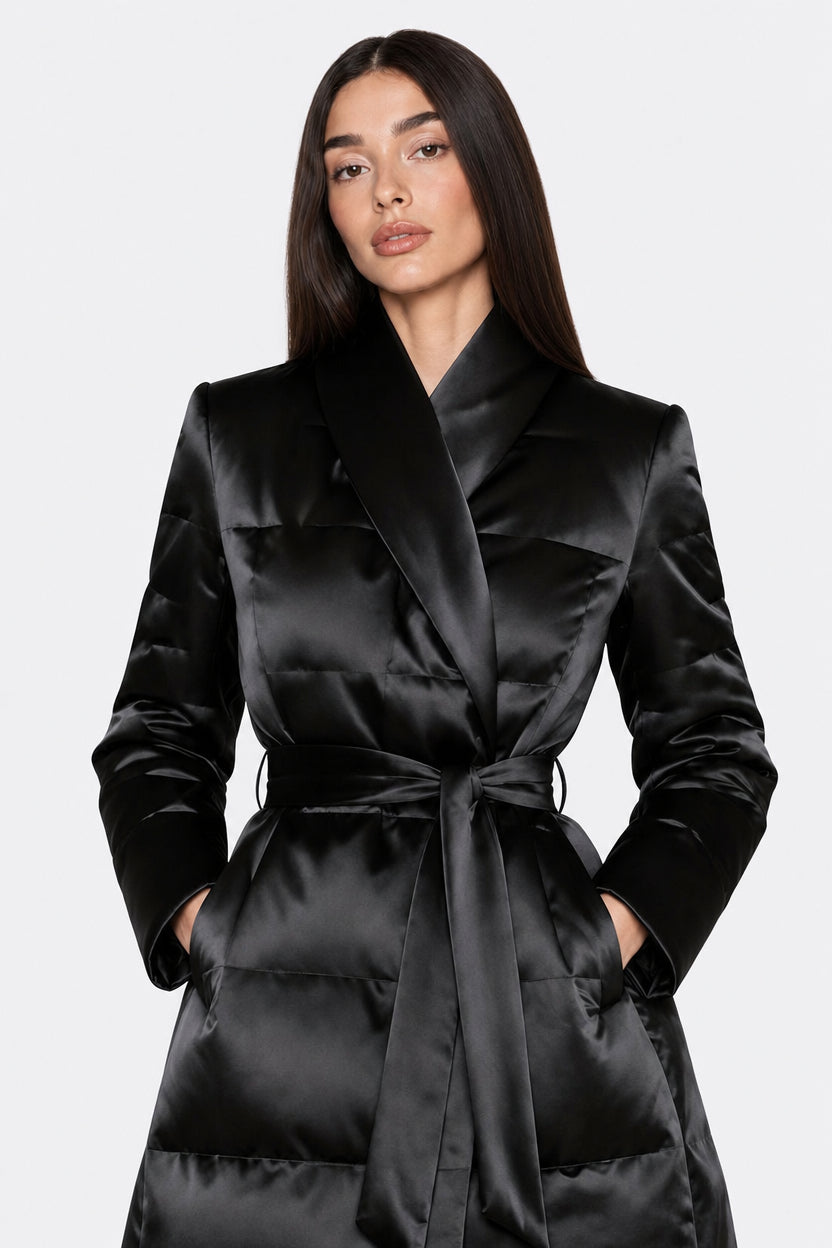 A woman modeling a black satin quilted puffer coat with a shawl collar and self-tie belt against a light gray studio background.