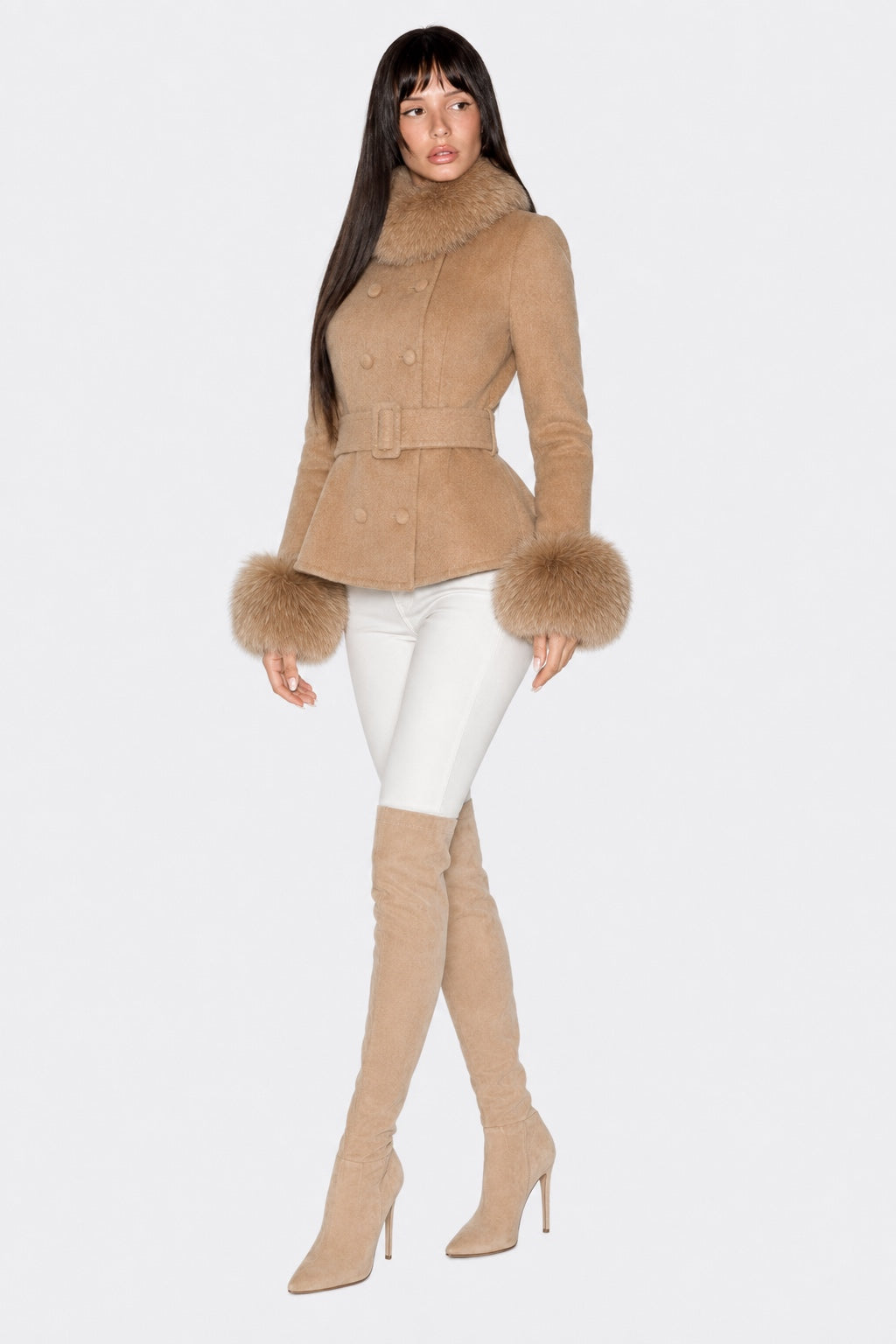 A woman modeling a camel-colored shearling-style coat with a matching belt and fluffy faux-fur trim at the collar and cuffs, paired with white pants and beige over-the-knee boots, against a light gray studio background.