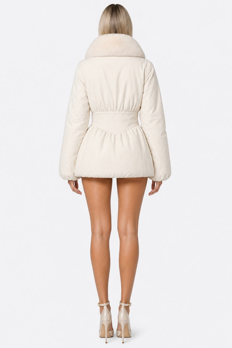 A woman shown from the back wearing a cream-colored belted puffer jacket with a fluffy collar, paired with beige high-heeled sandals, against a light gray studio background.