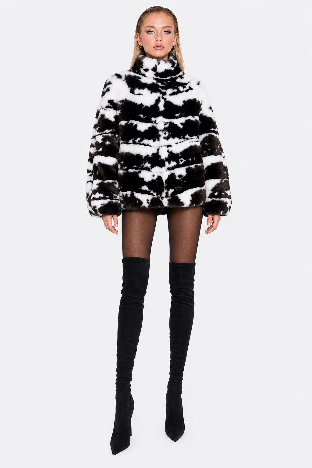 A model wearing a black and white tie-dye faux fur jacket with a high collar, paired with black tights and black over-the-knee boots, against a light gray background.